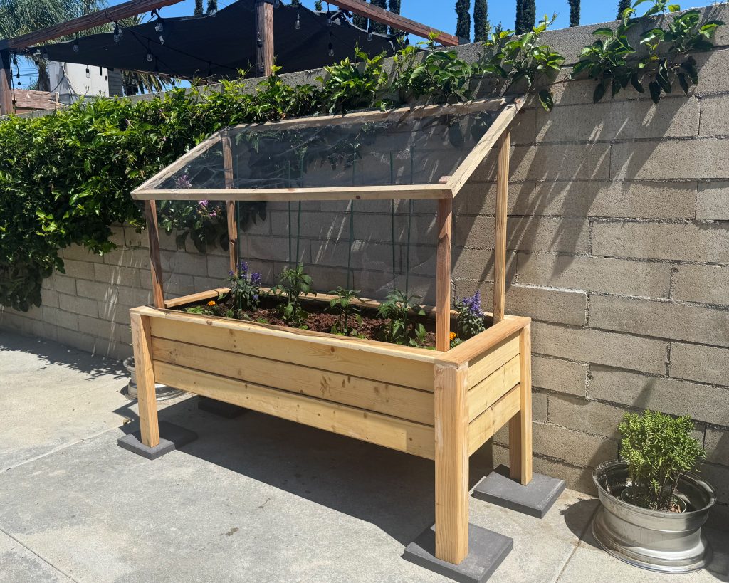 Angled view of a wooden raised planter with a propped-up screened lid, set on pavers beside a block wall with vines.